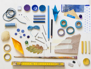 Assorted craft items on white table: blue pencils, tape rolls, thread, leaves, nails, paintbrush, yellow ruler, with vivid colors.