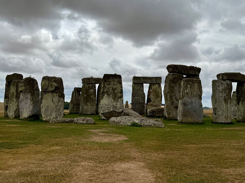 Photo of Stonehenge’s inner circle of standing stones on Salisbury Plain, with the Heel Stone visible in the distance directly center, under a grey sky with birds perched on the stones. 