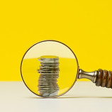 stack of coins and a wooden magnifier on a white table, yellow background. The concept of