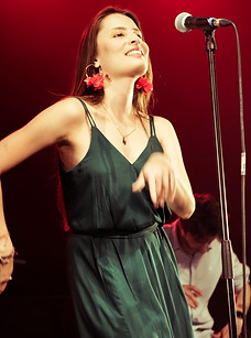 Une chanteuse aux longs cheveux bruns et une robe verte, avec des boucles d'oreilles rouges, se tient debout et danse en souriant devant un micro sur une scène éclairée de rouge, avec un musicien flou en arrière-plan.