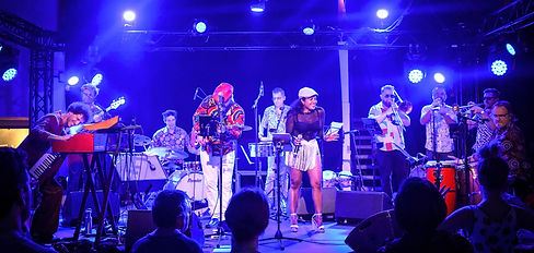 Sur scène, un groupe de musiciens énergiques joue de la musique boogaloo. Les membres sont habillés dans des styles rétro colorés et jouent des instruments variés comme le clavier, la basse, la batterie, les percussions, et des cuivres, avec des lumières de scène rougeoyantes au-dessus d'eux.