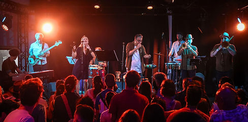 Une scène de concert avec un groupe de musiciens jouant pour un public debout, éclairés par des projecteurs, avec un mélange de percussions, de cuivres et de piano et basse électrique.