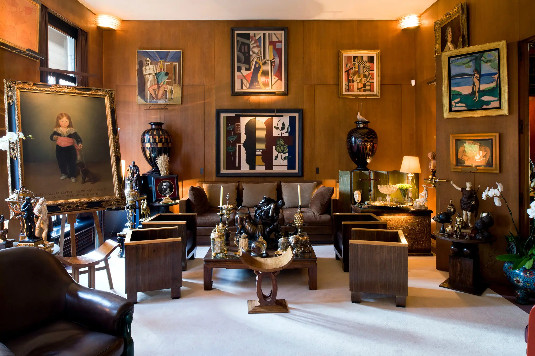 Living room as in the picture above with the same sofa, chairs, carved stool, pedestals with vases and chair in foreground that has been reupholstered in a dark leather. Bar cart has been traded out for art easel with painting of a boy. Coffee table has been changed to low wood one and holds a greater assortment of sculptures and art objects. The back wall now has four paintings, all done in various styles, and there are three more hung on the wall to the left. The rug has been removed as have all but two of the solid pillows on the sofa.