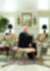 Nancy Pyne, appearing much older than in the previous photo, sits in the foreground on the same scrolled stool on top of the zebra hid rug. Same layout as the original room with hollyhock chintz on a sofa and chair, and the two same side chairs creating a seating group in front of the fireplace. The room is much smaller with a lower ceiling, white, painted, wide-plank wooden floors and windows on either side of the fireplace. The overall feeling is of a country cottage. A simple gilded mirror over the fireplace is flanked by two period sconces depicting eagles.