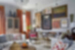 Large living room with white walls, floor to ceiling windows with orange silk drapes, and a traditional crystal chandelier hangs from the ceiling. Furnishings are all textural, some feeling rustic, some more refined like a marble topped, stepped coffee table with brass legs. Ethnic textiles along with carved wood and cane furnishings add a world traveller vibe and keep the space feeling casual. A collection of paintings, all different styles and subject matter and hung in a variety of frames grace one wall. Overall the room has a warm, eclectic, collected feel to it.