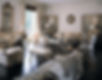 Large living room in a traditional style, divided into three separate seating areas. The overall feel is of a large manor or plantation home. One is flanked by antique painted screens with Sunburst clock on the wall between them and includes upholstered chairs and sofa along with a scroll leg bench. Antique wall mirror anchors another seating group which includes an upholstered sofa and side chairs. The final seating group includes chairs and sofa and another scroll leg bench, with a zebra print throw over the back of the sofa. Walls are all off-white with dentil crown molding and decorative half pillars throughout the room. The floor is a high shine dark brown with no floor coverings. The most striking design element is the large-scale black, white and grey chintz fabric that covers a number of the larger chairs and sofas. Several vases with large white floral arrangements are scattered around the room.