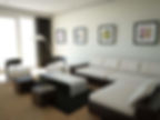 A living room with white walls, furniture two different colors, a dark brown and a white and all in a contemporary style. Furnishings include a low sectional and two chairs, a waterfall coffee table and two low ottomans. There is a very small rug under the coffee table in dark brown. An floor lamp with a matte silver finish arcs over the chairs. Curtains are dark brown. There are four square framed prints on the longest wall, each with a different flower on them which is the only color in the room. The feeling of the room is sterile with no personal touches.