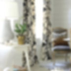 Corner of a living space with  floor to ceiling drapes in Pyne Hollyhock pattern.  Adjacent chair is a traditional style with grey upholstery and leopard print pillow.  In the left foreground is the arm of a sofa and side table with a lamp, potted plant and some decorative beads.  The walls are a white shiplap and the overall style is modern farmhouse.  Design credit in lower right corner to Sherry Hart Design.