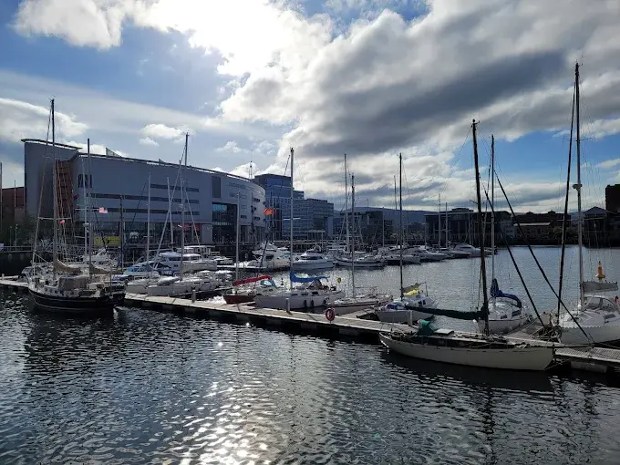 Daytime view of Belfast marina with yachts docked in calm water, modern waterfront buildings in the background, bright sky with scattered clouds, wide landscape.