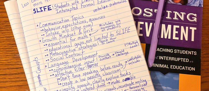 Notebook with handwritten notes on educational strategies for SIFE, next to a book titled "Boosting Achievement." Purple pen on top, wooden table.