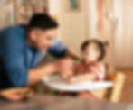 A father feeding his baby girl green beans in a high chair, smiling during mealtime as part of a healthy eating routine.
