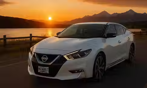 White Nissan Maxima parked by lake at sunset with mountains in background.