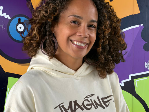 A picture of Brenda Liz Henry in front of a graffiti mural honoring Hip Hop. Brenda is wearing a beige hoodie, has curly brown hair, and is wearing hoop earrings.