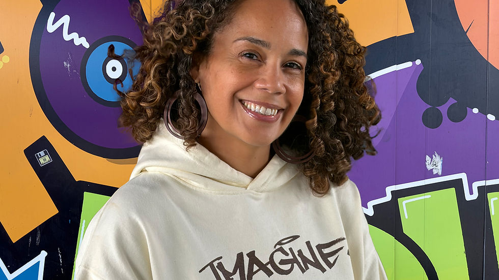 A picture of Brenda Liz Henry in front of a graffiti mural honoring Hip Hop. Brenda is wearing a beige hoodie, has curly brown hair, and is wearing hoop earrings.