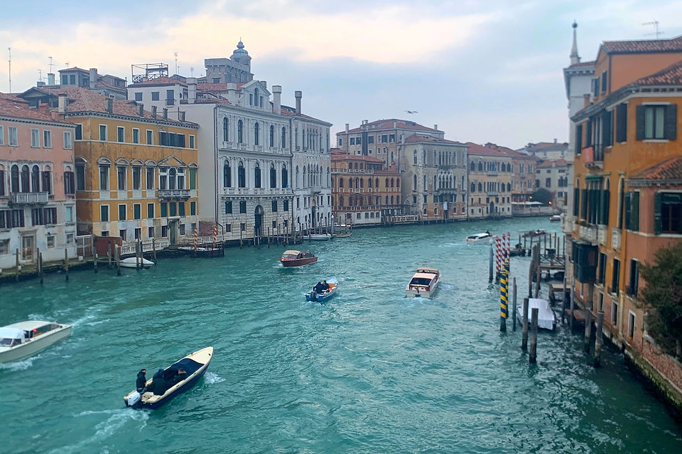 A view over the Grand Canal in Venice, Italy. Boats drive through the deep turquoise waters on an overcast day. Yellow, pink, and beige palazzos and buildings flank the river. Photo by Darcy and Nathaniel