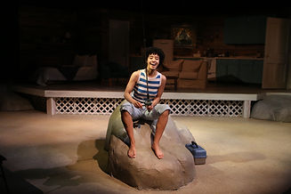 Production photo from Last Summer at Bluefish Cove of an actor sitting barefoot on a rock and fishing