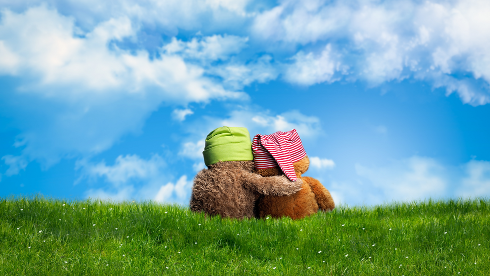 Two teddy bears sit in the grass with their backs facing front and the left bear has its arm around the other.