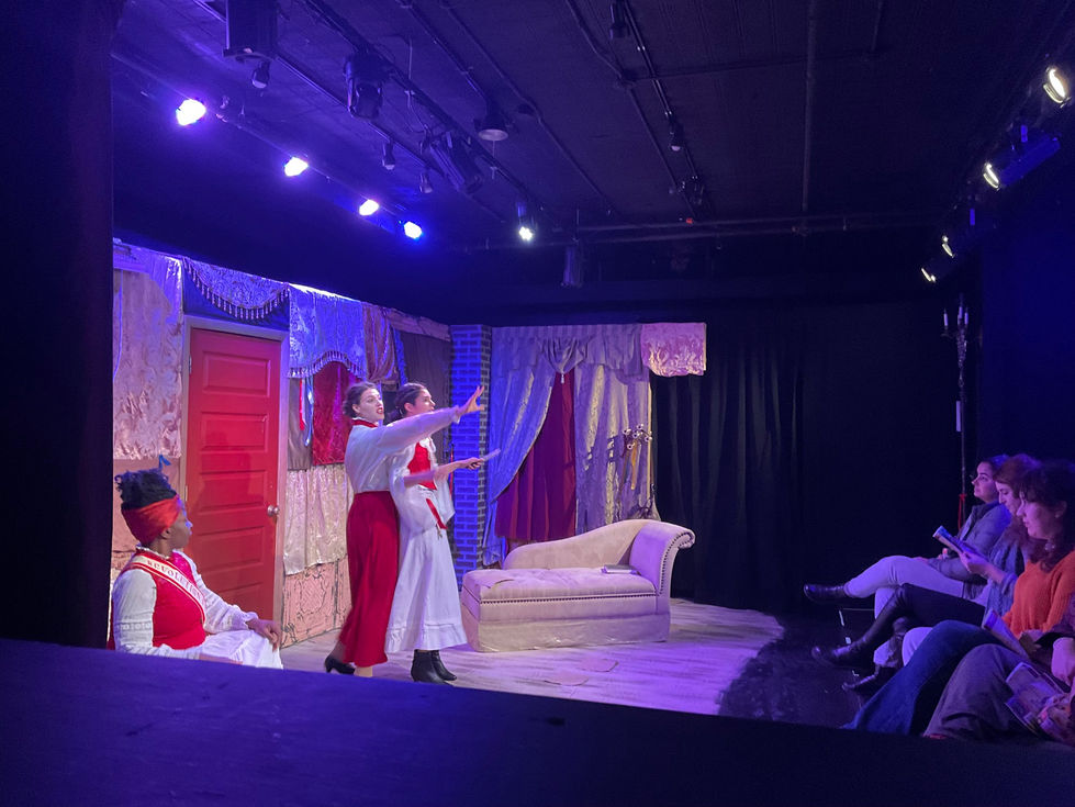 Mid-Production photograph of The Revolutionists by Lauren Gunderson performed at Madison Street Theater. Actors are in white and red period costumes and the audience is seen across from them in the intimately staged blackbox space. Olympe de Gouges is drawing an image with her words between the cast and the first row of the audience.
