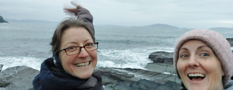 Two women in hats and coats laughing in front of a camera, behind is the Atlantic ocean. 