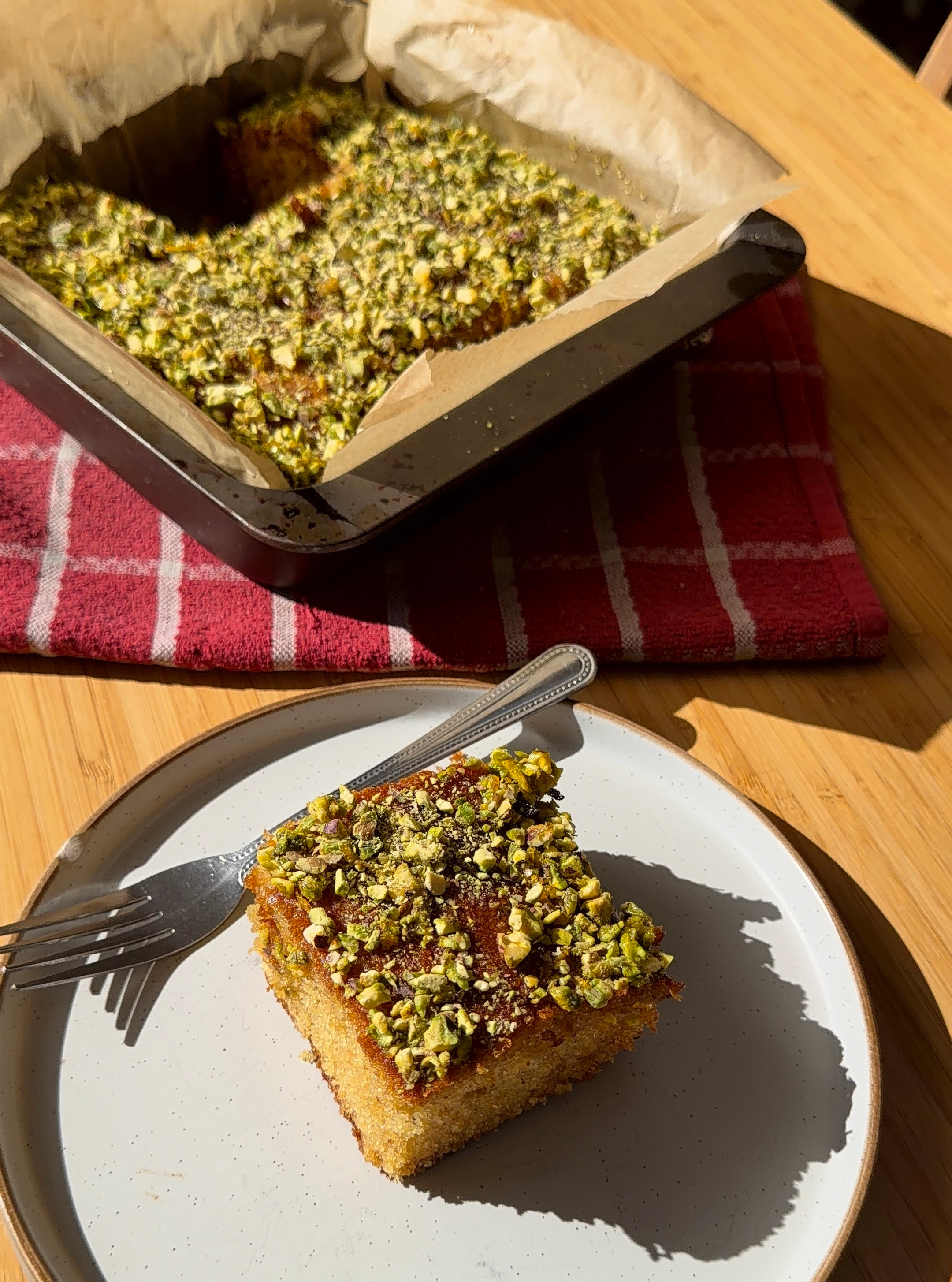 Pistachio-topped cake in a baking pan and a plated slice with fork, lit by warm sunlight on a wooden table.