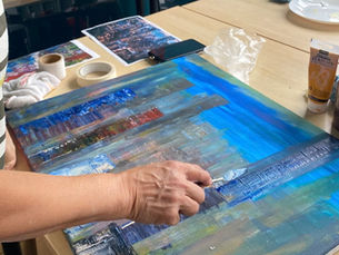 A hand holding a palette knife adding colour to a skyscraper city scene painting