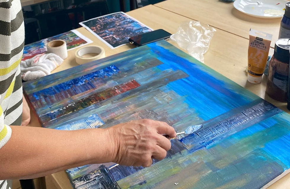 A hand holding a palette knife adding colour to a skyscraper city scene painting