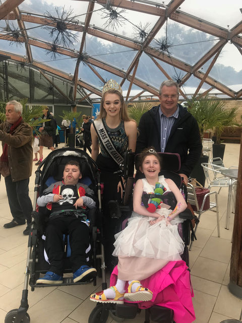 A man and woman stand behind a boy and girl in wheelchairs. The woman is dressed in a pageantry outfit and a tiara. The boy is dressed as a pirate and the girl as a princess.