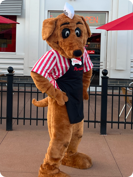 Mascot dog in a striped shirt and apron posing outside a restaurant.