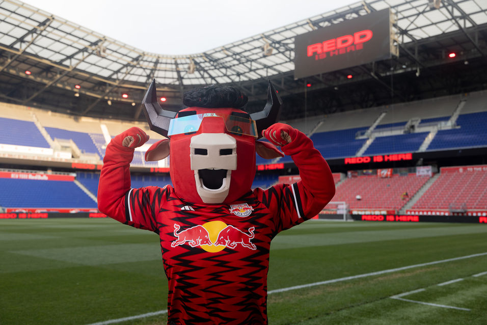 Red bull Mascot flexing in a stadium setting with stands and screens in the background.