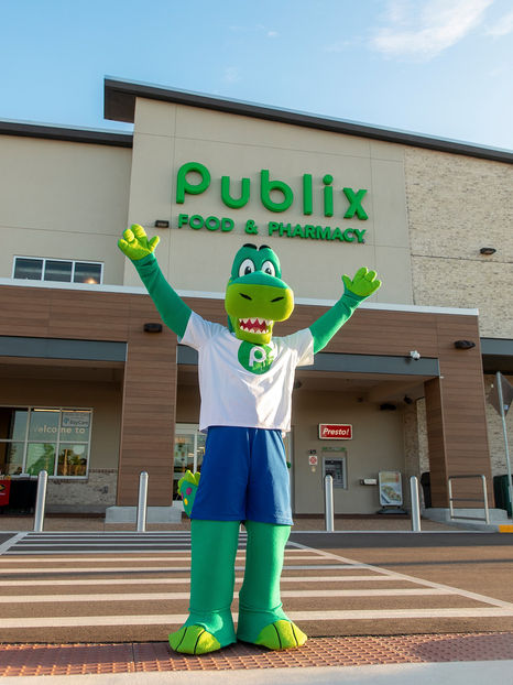 Publix alligator mascot standing with raised arms in front of a Publix store.