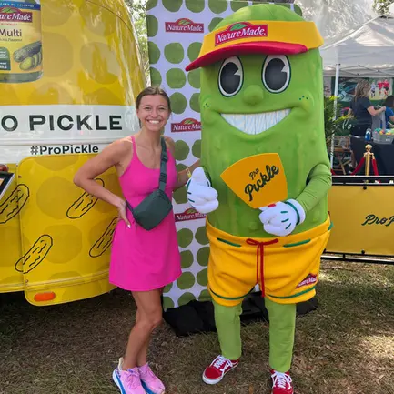 Woman posing with a Nature Made pickle mascot at an outdoor event.