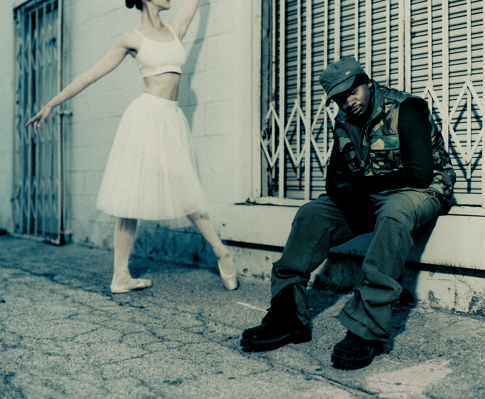 Woman in white ballet outfit poses beside seated man in camo vest and cap outside a shuttered storefront.