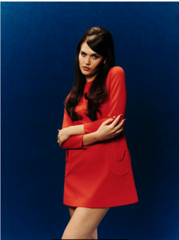 Woman in a red dress stands with arms crossed against a dark blue background, giving a serious, poised look.