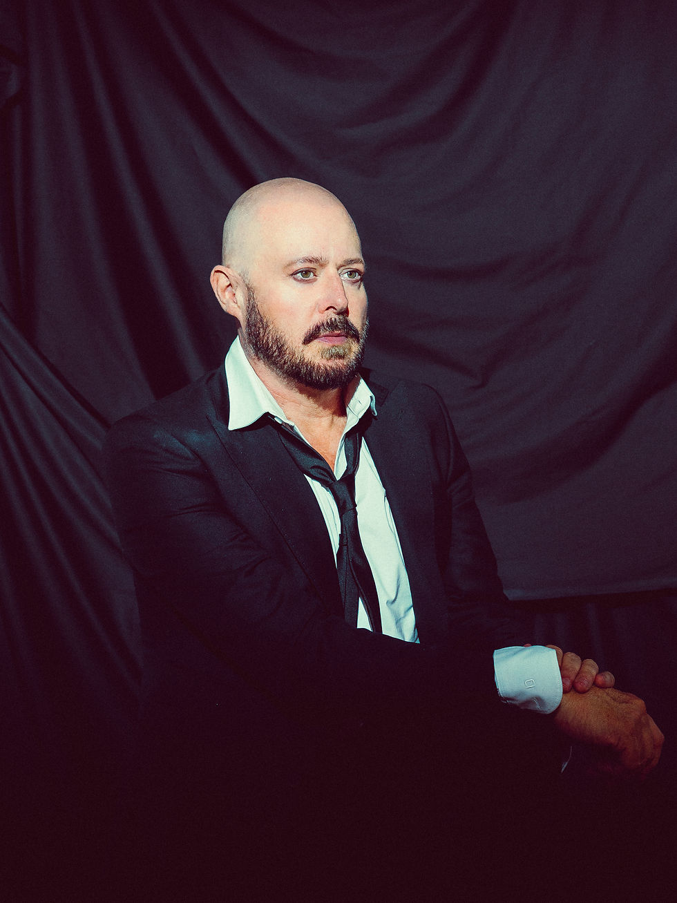 Bald bearded man in a black suit and loosened tie sits against a dark draped backdrop, looking serious and reflective.