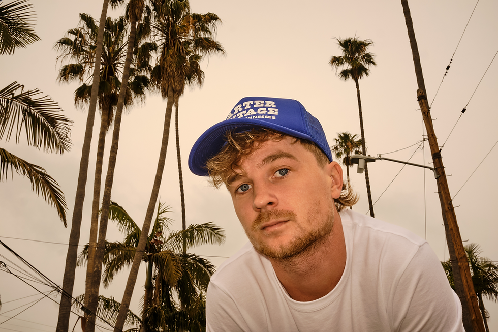 Close-up of man in blue Carter Vintage Tennessee cap and white shirt, looking at camera with palm trees and power lines behind.