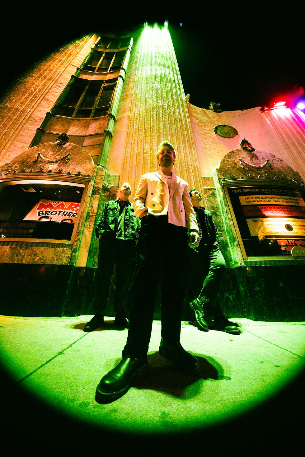 Three men pose under neon green and gold lights at a nighttime museum entrance, with show signs visible in the windows.