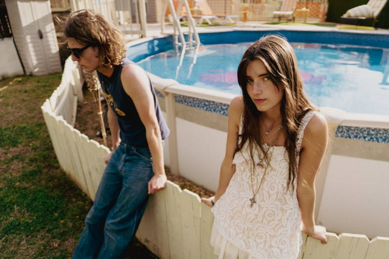 Two young people lean on a white fence by a blue backyard pool, one in sunglasses, one in a white dress, in warm sunlight.