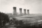 A black and white image shows a large field, with the edge of trees along the bottom left side. In the center distance, four massive cooling towers stand out against the landscape.