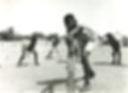 A black and white photograph shows five or six children playing cricket. One child stands in front of the other four, holding out a cricket bat as if they have just taken a swing.