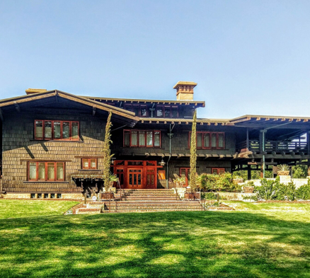A color photograph shows the front exterior of the Gamble House. The lawn is a lush green, with decorative plants near the front of the home. The sky is clear blue.
