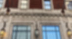 Image shows a cropped section of the side of a building, the bottom half is white decorative marble around two windows, the top is red brick. At each side, there is a small round carved portrait.