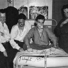 A black and white photograph shows four men. The center man sits on a hospital bed, directly facing the camera with a slight smile. Two men stand to his left, one who faces the camera with a cigarette hanging from his mouth, and one who is laughing but looking towards the floor. Another man stands to the right of the hospital bed, with a serious, moody expression, looking towards the floor. Two pinup pictures of attractive women hang behind the men on the wall. All four men have tube pedicles that stretch down from their noses.