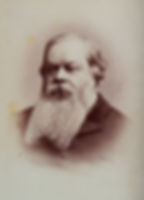 An old sepia photograph shows a middle-aged white man with a long beard. His hair is parted on the left side and is ear length, he looks off to the side, unsmiling. 