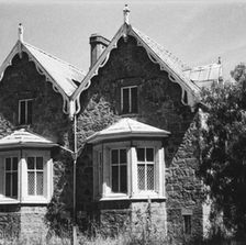 A black and white image shows the side of a house, flanked by two trees. The house is a Gothic Revival style, and the design appears to be mirror-imaged down the center, with identical pairs of windows on both sides. 