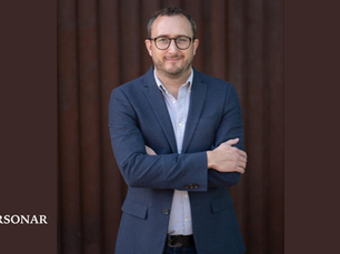 Tony Page, press photo, with short hair, glasses, and light stubble stands with his arms crossed, wearing a blue blazer over a light shirt in front of a dark wooden background. The PERSONAR logo appears in the lower left corner.