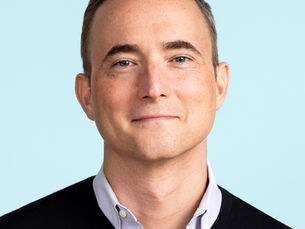 Eric Stevens wearing a black sweater over a white collared shirt, against a light blue background.