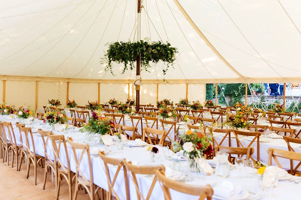 Bentwood cross back chairs are positioned at long trestle tables inside a Peacock and Bow pole and canvas marquee. The tables are dressed with white linen and floral arrangements and foliage cascades from a flower ring hanging high in the canopy.