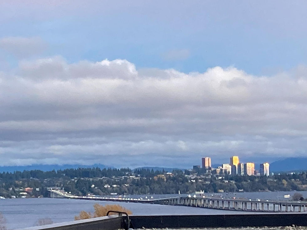 Cityscape with a bridge over water, leading to buildings lit by golden sunlight. Cloudy sky contrasts with blue patches, creating a serene mood.