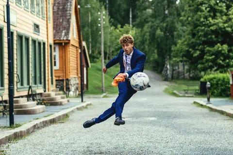 Ung mann i blå dress som hoppe opp og skyter en fotball i et bymiljø,