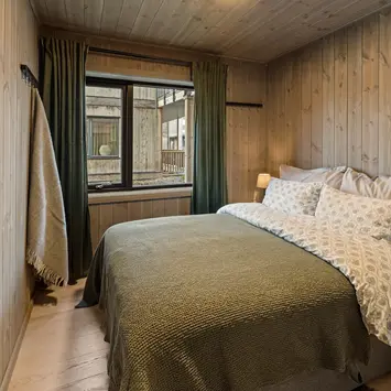 Soverom med dobbeltseng, trepanel, vindu med grønne gardiner og grønt sengeteppe.